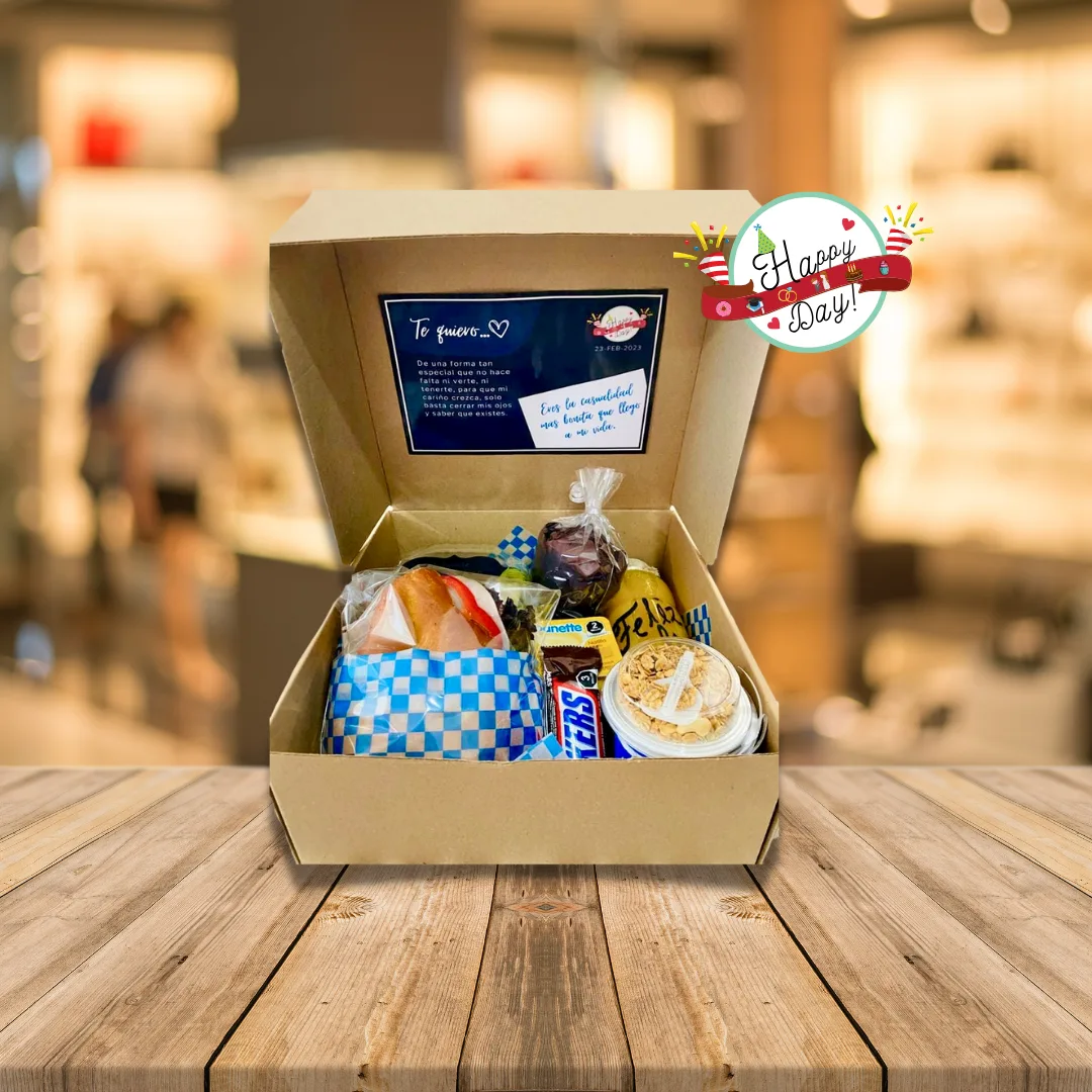 Caja de cartón abierta con cuernito, mix frutal, natilla, chocolate, yogurt y una tarjeta de dedicatoria.